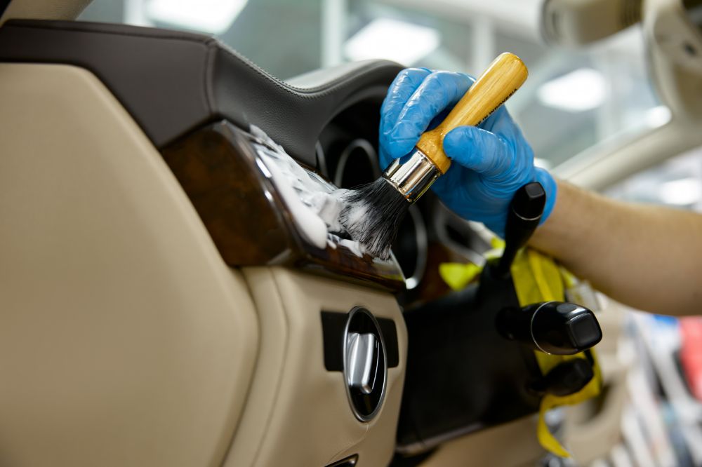 Person durchführt Autoaufbereitung in Oldenburg, reinigt sorgfältig das Innere eines Autos mit einer weichen Bürste und Schaum auf der beigen Lederinnenverkleidung.